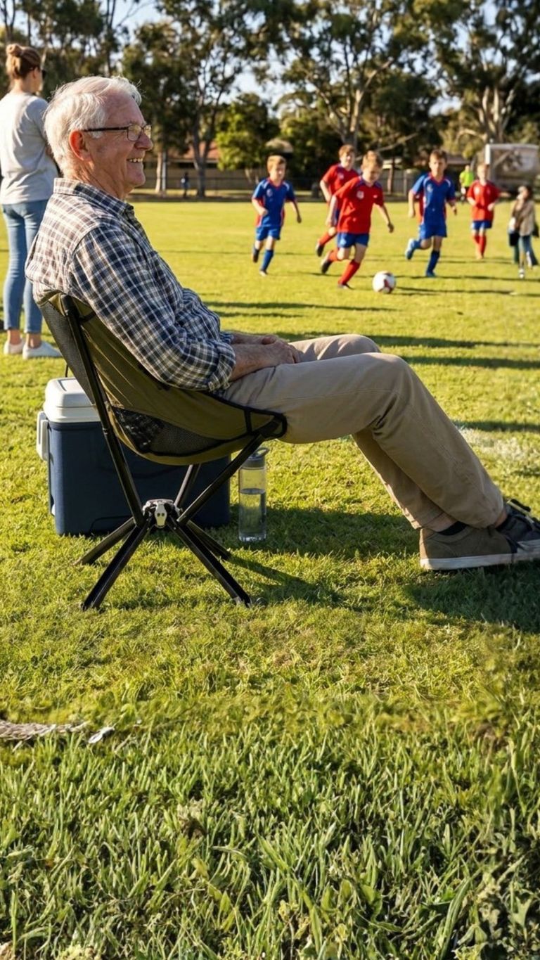 An elderly man sits in a portable chair on a sunny grass field, smiling as he watches young children play soccer in the background, suitable for lifestyle or family-oriented content.